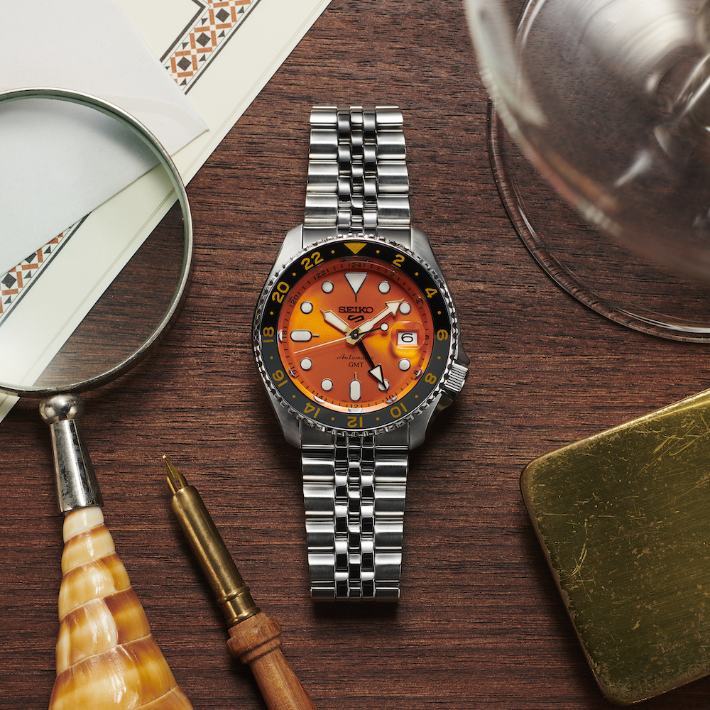 Seiko Sports 5 GMT in orange on a table next to a magnifying glass